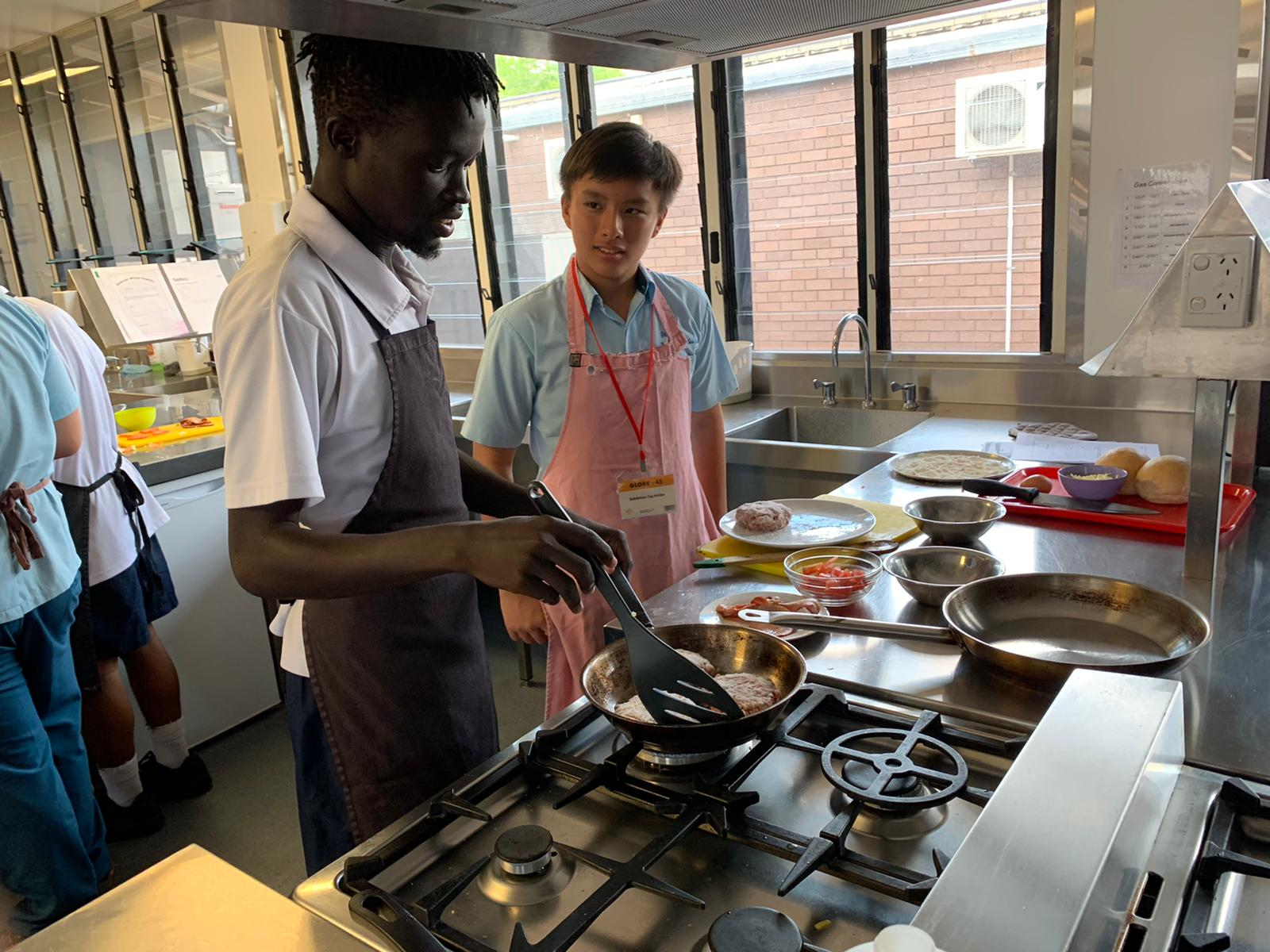 Students wearing aprons prepare food at cooking stations in a commercial kitchen during a hands-on culinary lesson.